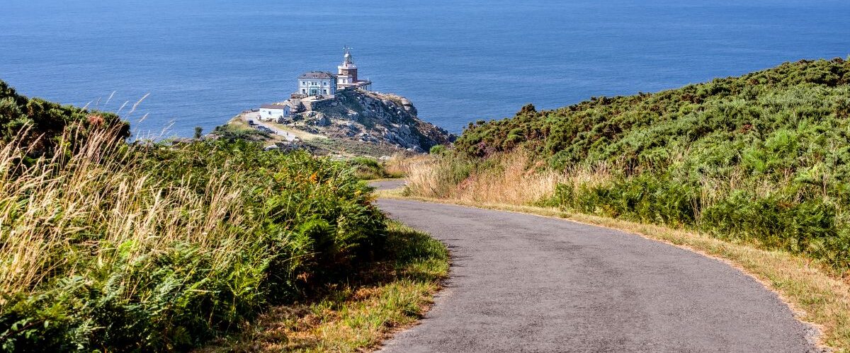 Spain, Finisterre: Famous lighthouse on rocky cliff with road, seascape, ocean sea, skyline and blue sky in the background. It is located on a rock-bound peninsula and was known as end of the world.